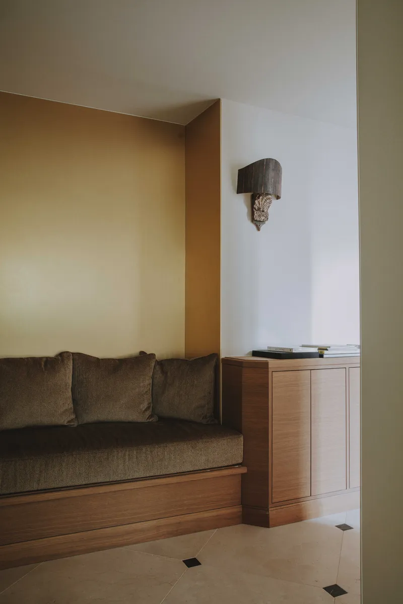 Banquette d’accueil en niche laiton, velours et merisier, éclairée par la lumière du jardin.