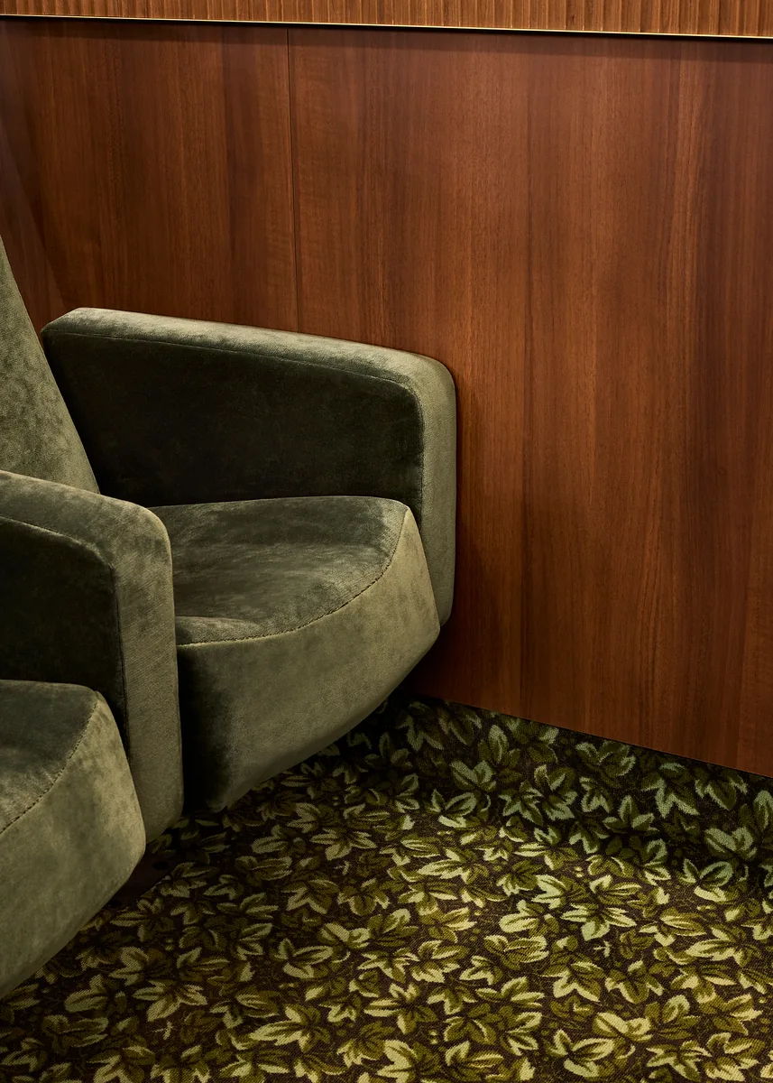Detail of a cinema seat and mahogany panelling, with a brass reveal and foliage carpet.