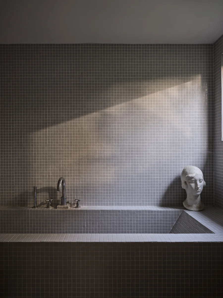Baignoire maçonnée en mosaïque grise, rai de lumière et tête en plâtre.