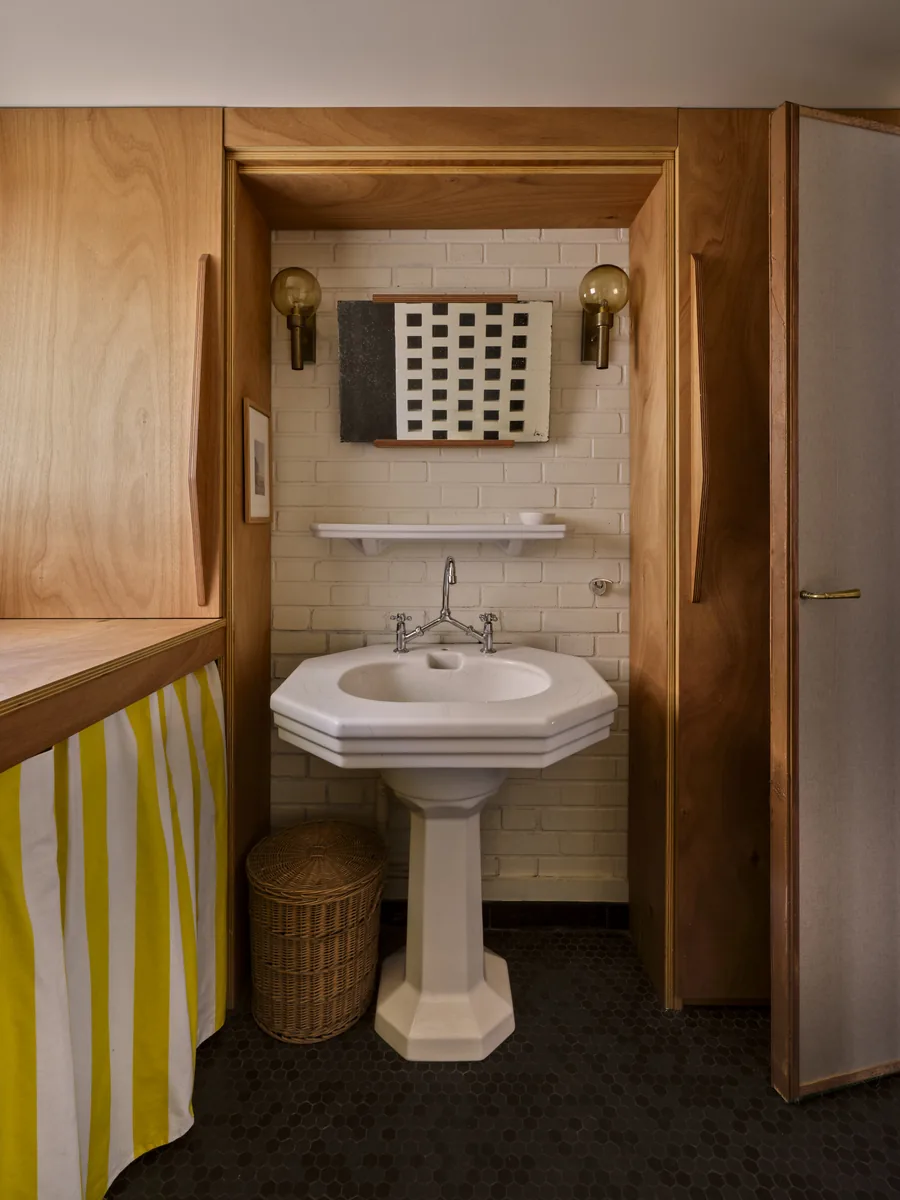Salle d'eau avec lavabo colonne, brique peinte, sol en tomettes noires, appliques en laiton et rideau rayé jaune.